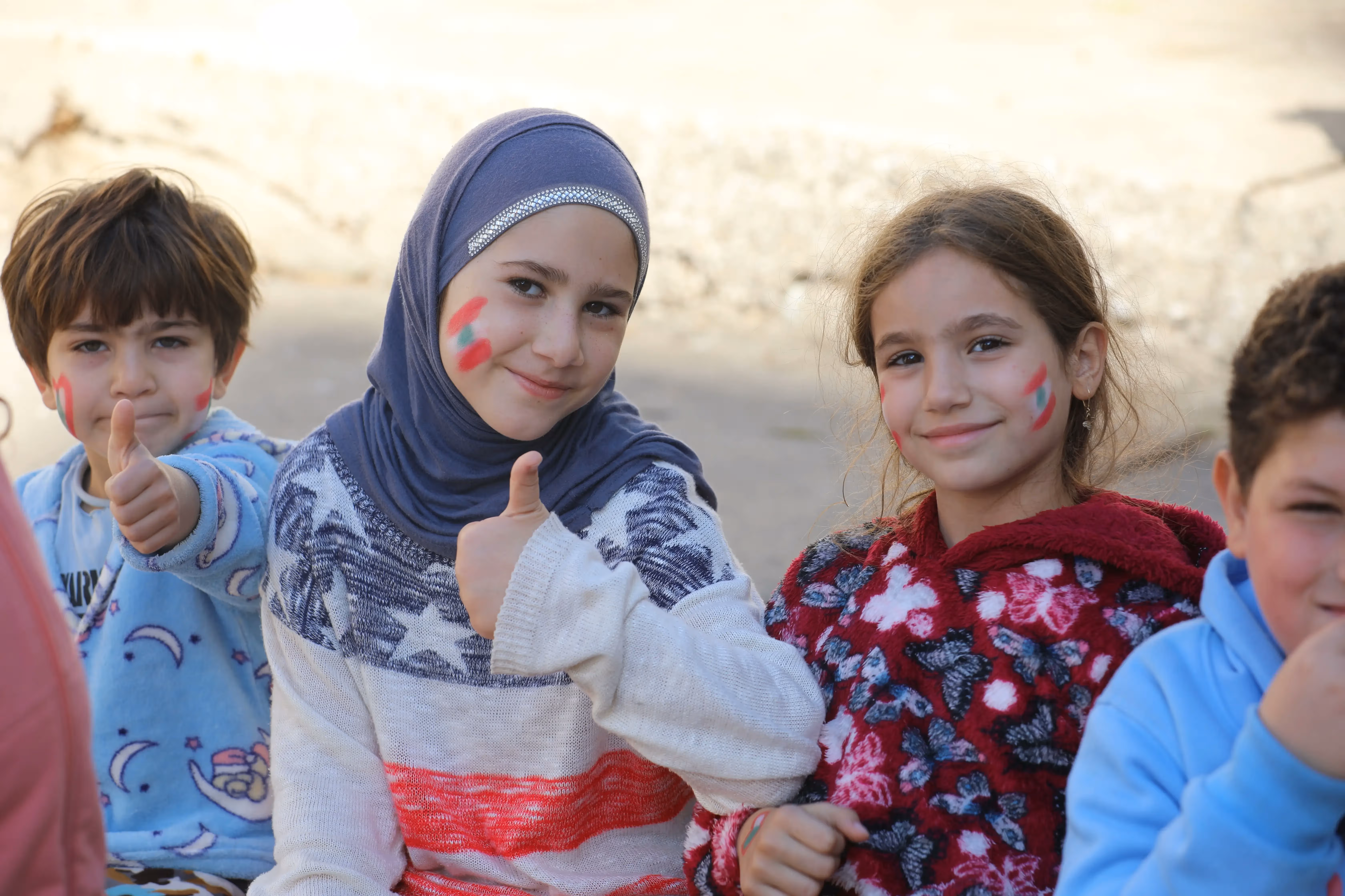 Group of smiling children with the flag of Lebanon painted on their cheeks giving thumbs-up gestures.